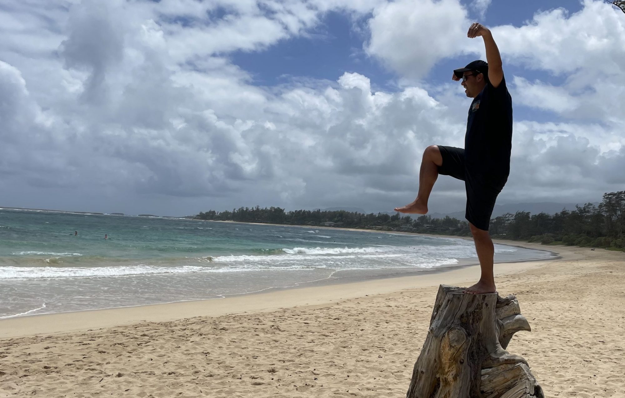 Anthony balancing on a driftwood stump on an empty beach, ocean and clouds behind him