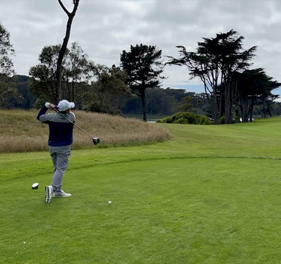 Golfer mid-swing on a tee box lined with cypress trees