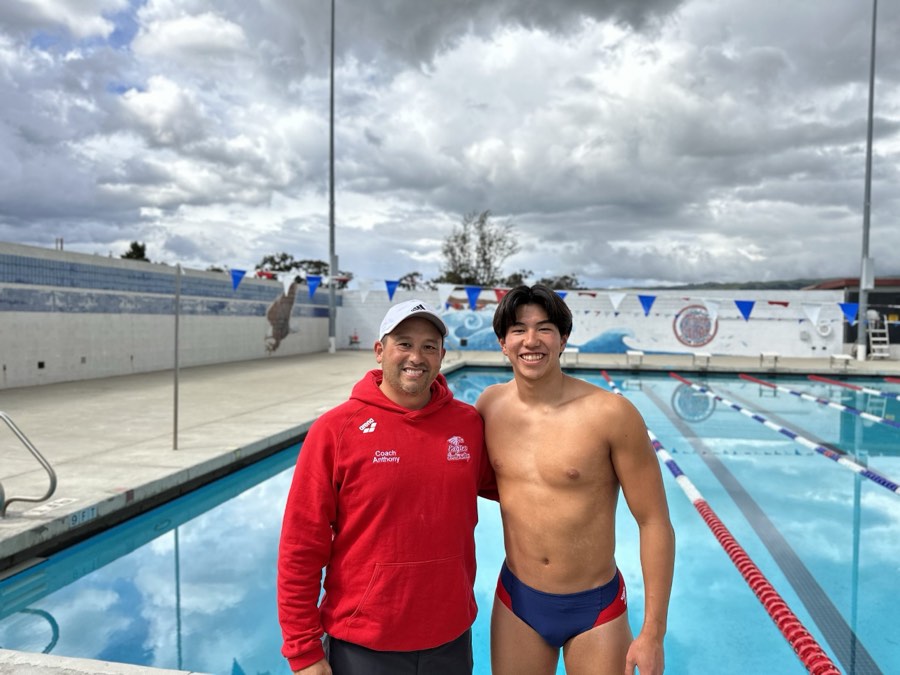 Coach in a red jacket standing with a swimmer beside an outdoor competition pool